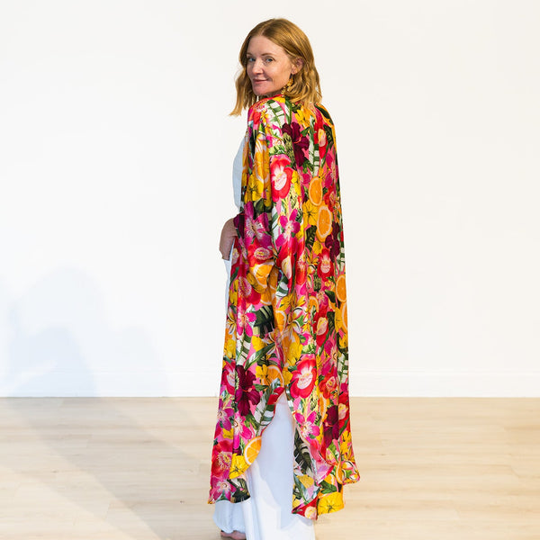 A woman stands barefoot on a light wood floor against a white wall, wearing a long, flowing floral robe or drape in bright pink, yellow, orange, red, and green tones over white pants.