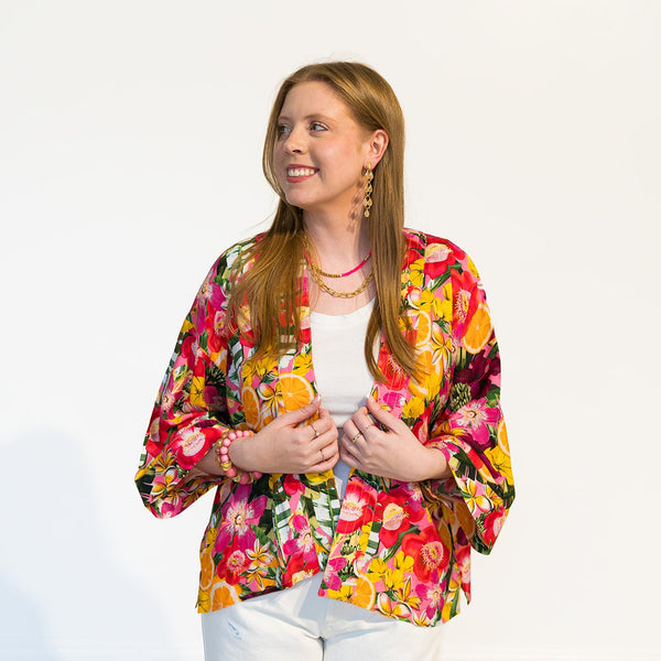 A woman with long light brown hair stands against a plain white wall wearing a vibrant, multicolored floral kimono-style jacket over a white top and white pants.