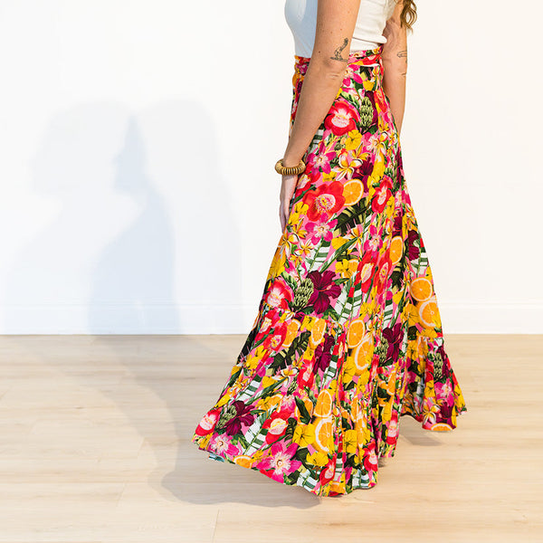 A woman wearing a long, flowing floral maxi skirt in a bright studio setting with a white wall and light wood floor. The skirt features a dense multicolor floral print with prominent pink, yellow, orange, and red tones.