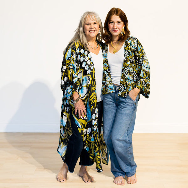 Two women wearing floral kimono jackets standing on a light wooden floor with a white background