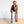 A woman stands barefoot on a light wood floor against a white wall, wearing a long open-front robe with a vibrant multicolor retro concentric circle pattern over a black top and white pants.