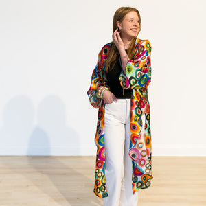 A woman stands barefoot on a light wood floor against a white wall, wearing a long open-front robe with a vibrant multicolor retro concentric circle pattern over a black top and white pants.