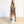 A woman stands barefoot in a minimalist studio with a white wall and light wood floor, wearing a long, flowing kaftan-style robe with a vibrant multicolor circular pattern over white pants.