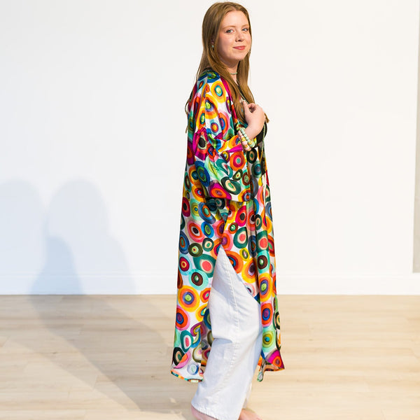 A woman stands barefoot in a minimalist studio with a white wall and light wood floor, wearing a long, flowing kaftan-style robe with a vibrant multicolor circular pattern over white pants.