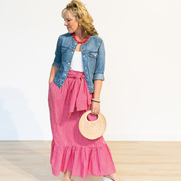 Woman wearing a denim jacket, white top, and pink ruffled skirt with a beige bag on a plain background