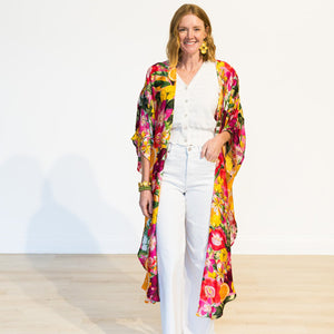 A woman walks on a runway wearing a vibrant, multicolored floral kimono jacket over a white button-up top and white wide-leg pants, set against a plain white studio background.