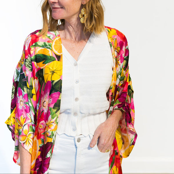 A woman with blonde hair is wearing a white button-up top and white pants, styled with a vibrant, multicolored tropical floral kimono jacket. She is standing against a plain white background.