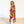 A woman stands barefoot on a light wood floor against a white wall, wearing a long, flowing floral robe or drape in bright pink, yellow, orange, red, and green tones over white pants.