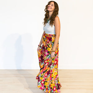 A woman wearing a white sleeveless top and a vibrant, multicolored floral maxi skirt stands in a studio setting with a plain white wall and light wood floor.