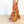 A woman wearing a long, flowing floral maxi skirt in a bright studio setting with a white wall and light wood floor. The skirt features a dense multicolor floral print with prominent pink, yellow, orange, and red tones.